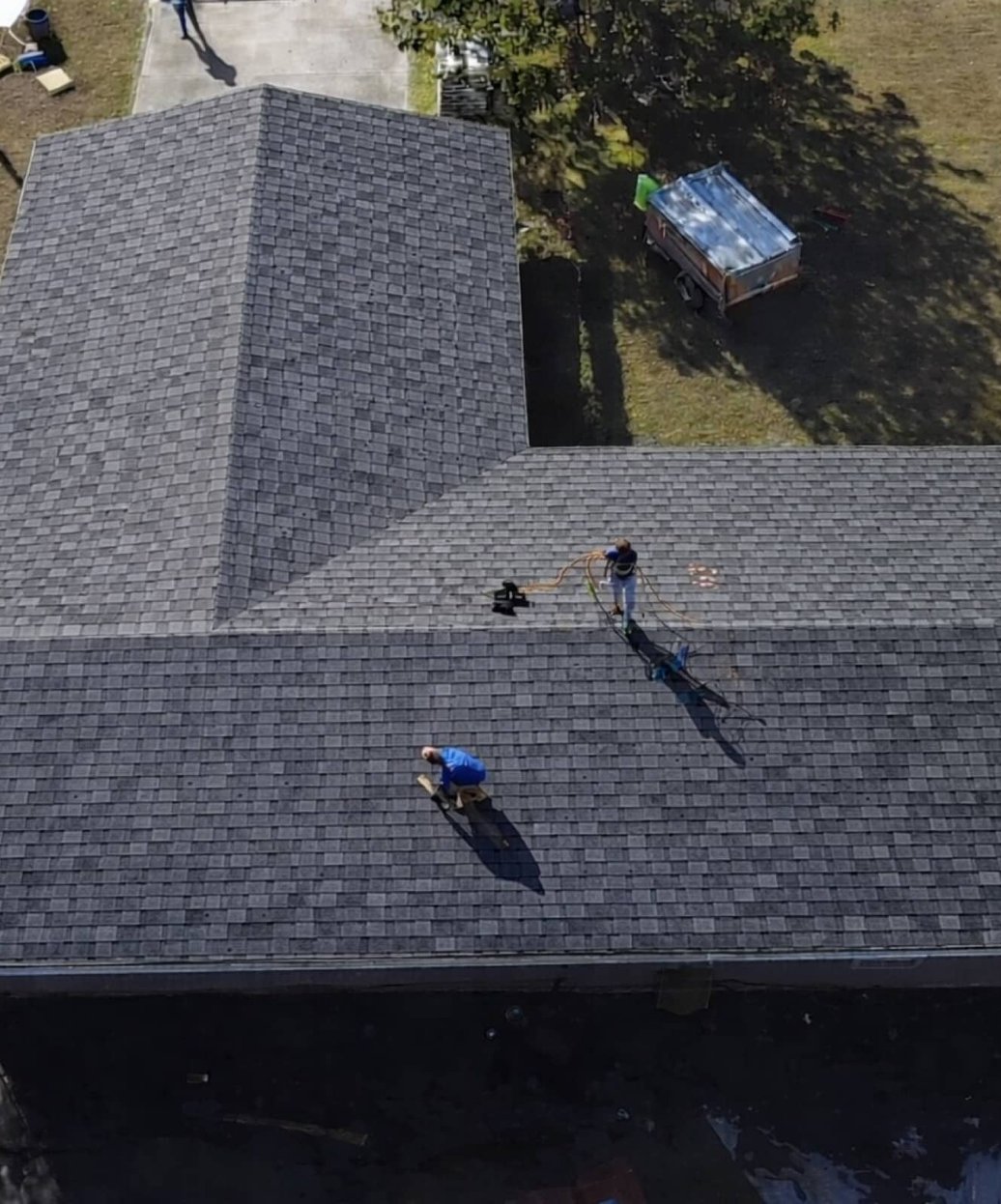 Two roofers working on a dark grey shingle roof, seen from a drone above an L-shaped house