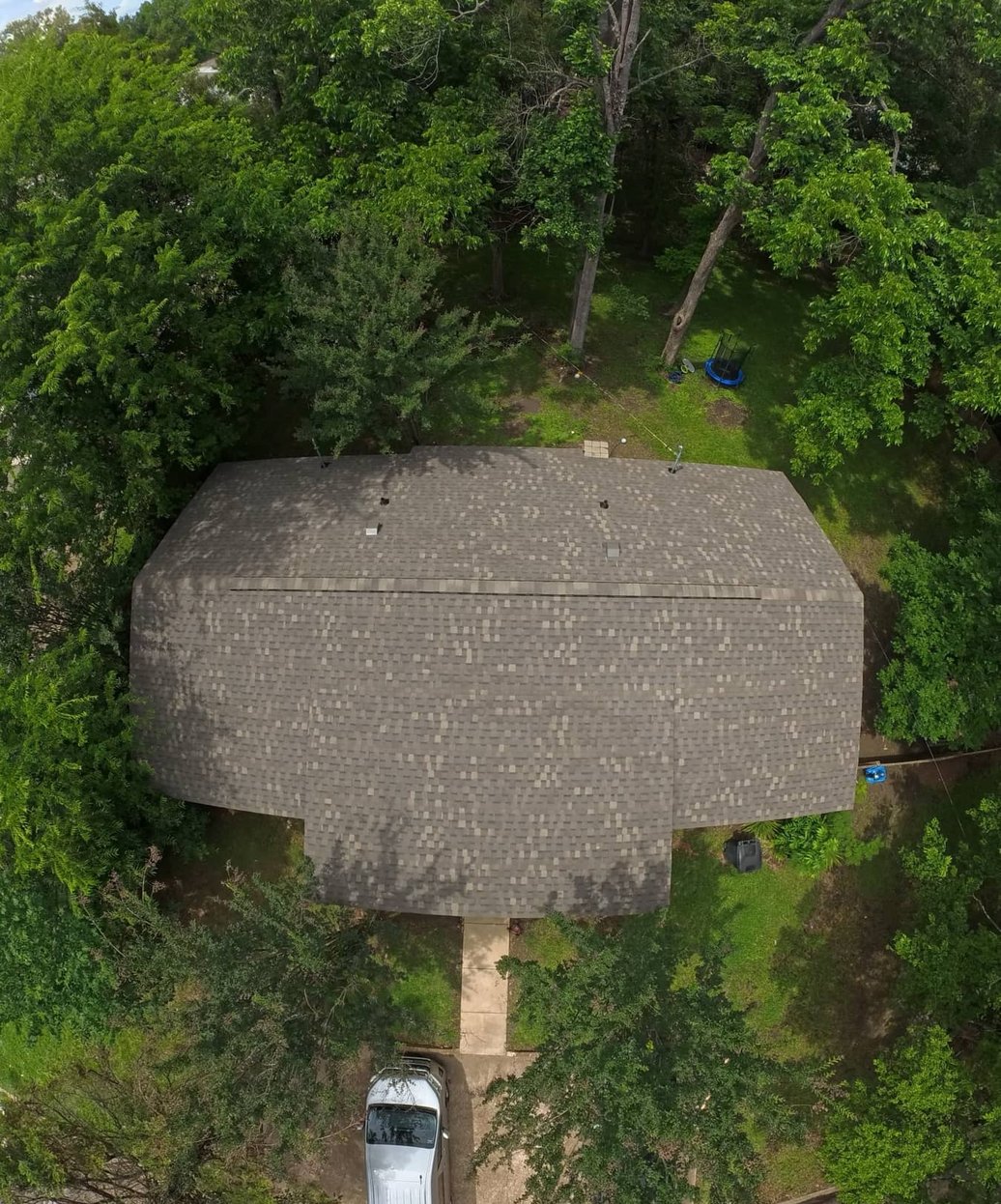 Aerial view of a grey shingle roof on a home tucked among dense trees, with a white truck in the driveway