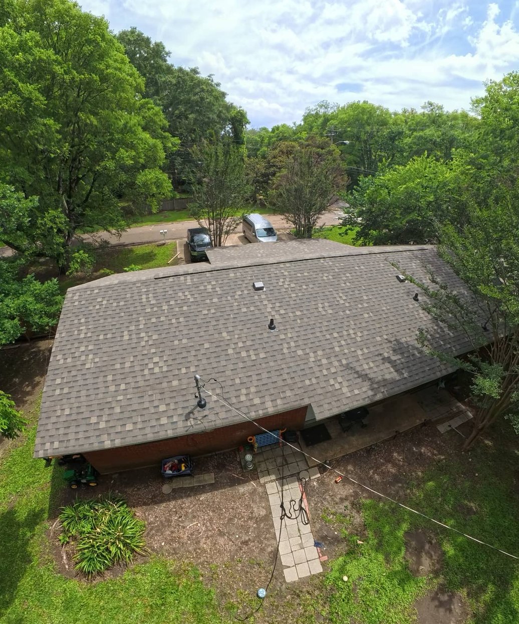 Drone view of a finished grey shingle roof on a single-story home with trees and a driveway behind it