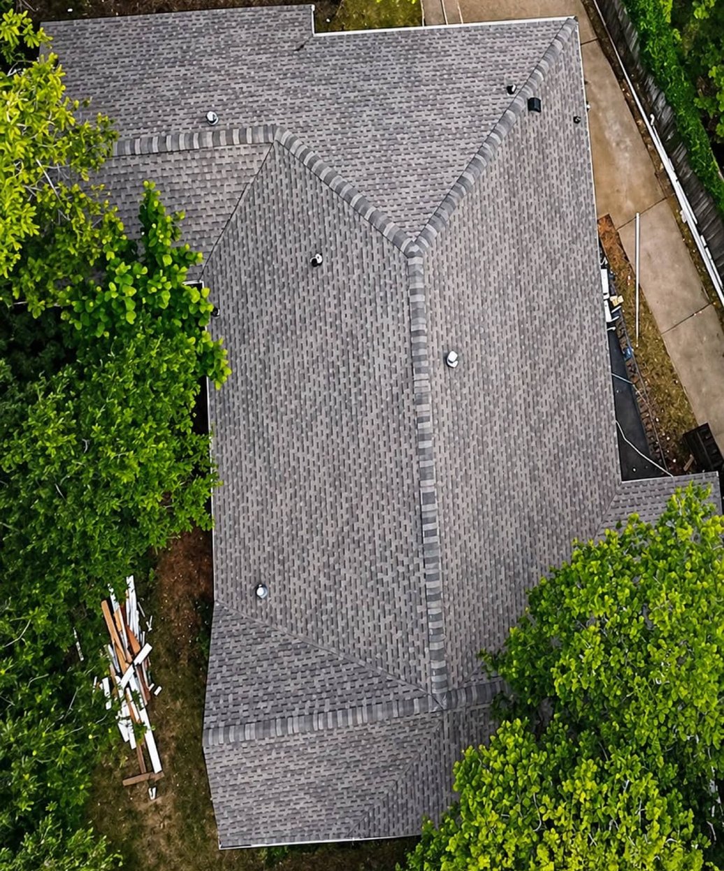 Top-down view of a grey architectural shingle roof with hips and valleys among green trees