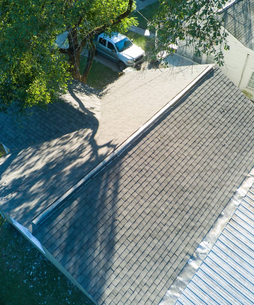 Aerial view of a grey shingle roof beside a metal roof, with a pickup parked under a large tree